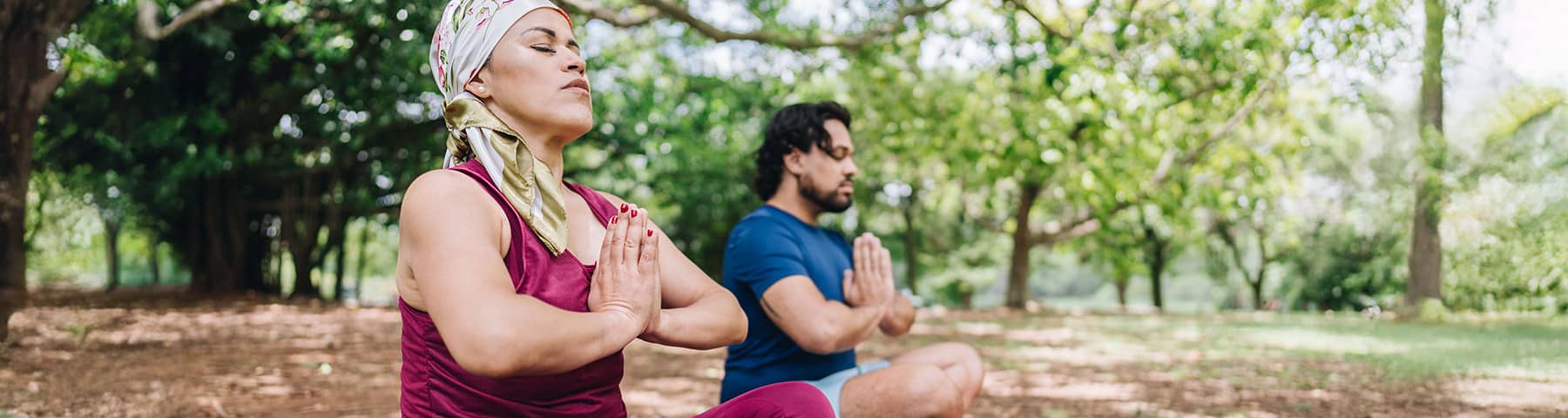 Two people meditating in a park, seated cross-legged with hands in prayer position.