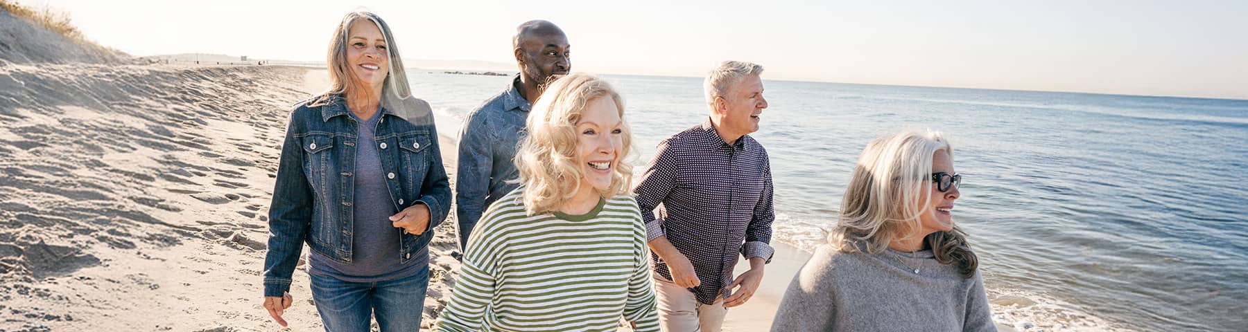 Five older adults smiling and walking together along a sunny beach with the ocean in the background.