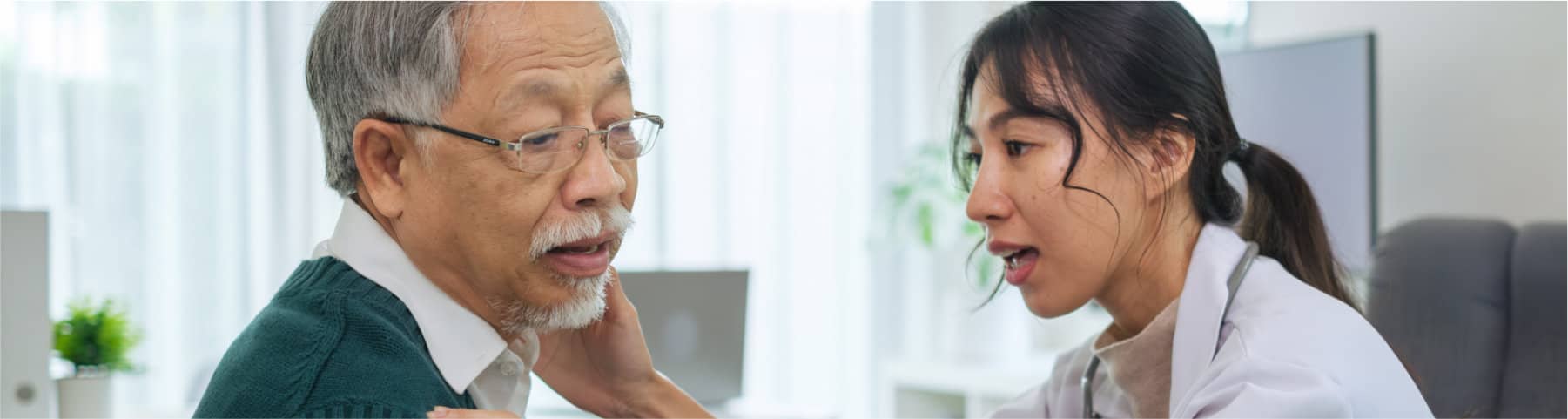 A female doctor in a white coat examines the neck of an older male patient in a brightly lit office.