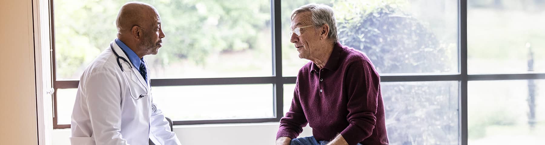 A doctor speaks with an older male patient in a well-lit medical office.