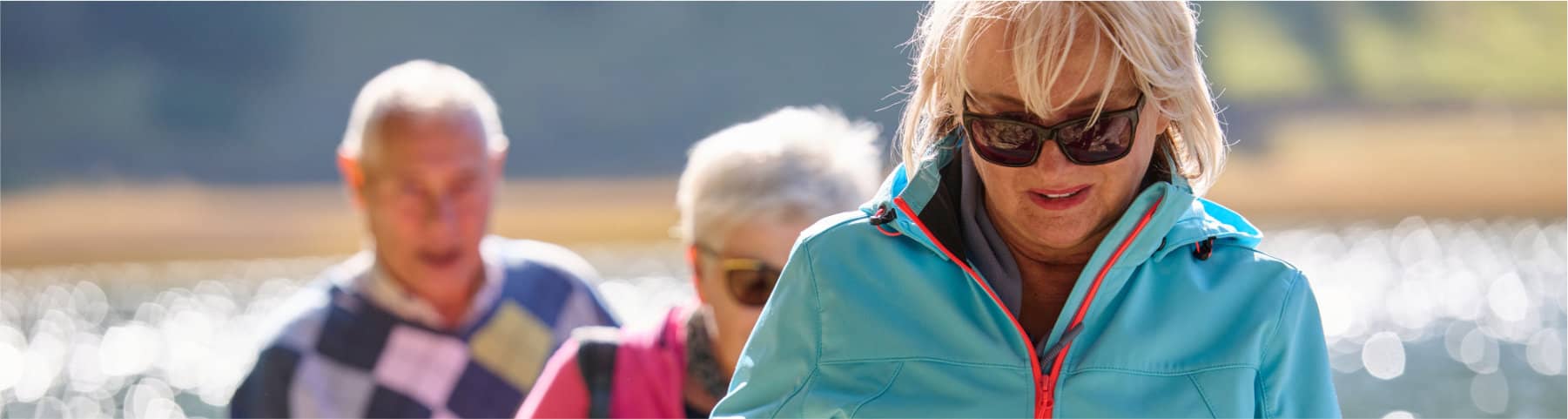 An older woman wearing sunglasses and a blue jacket walks outdoors, with two other seniors visible in the blurred background.