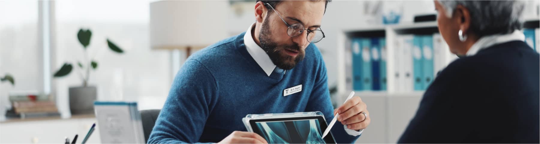 A male doctor with glasses reviews an X-ray image on a tablet while consulting with an elderly female patient.