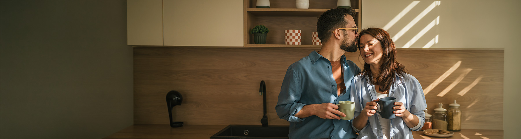 A smiling couple holds coffee mugs in a sunlit modern kitchen, sharing an affectionate moment.