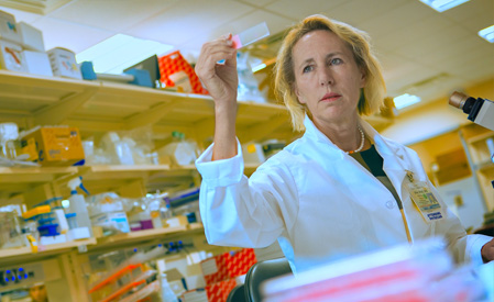A female scientist in a white lab coat examines a small glass slide in a well-lit laboratory. Shelves with lab supplies and a microscope are visible in the background.