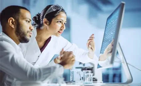 Two scientists in white lab coats collaborate in a laboratory, reviewing data displayed on a computer monitor, with a microscope visible in the foreground.