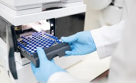 A close-up of gloved hands in blue nitrile gloves loading a tray of small sample vials into an automated laboratory instrument or autosampler.