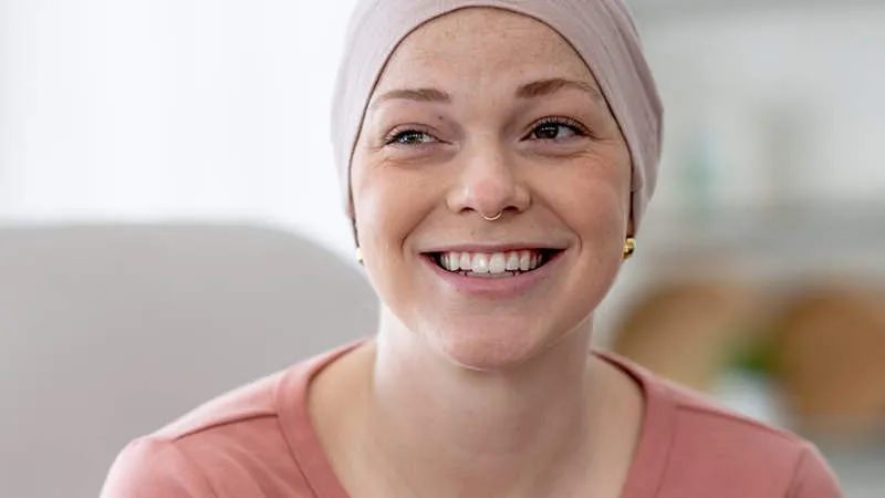 A young woman with freckles wearing a pink head scarf and a pink t-shirt, smiling warmly in a bright, softly blurred indoor setting.