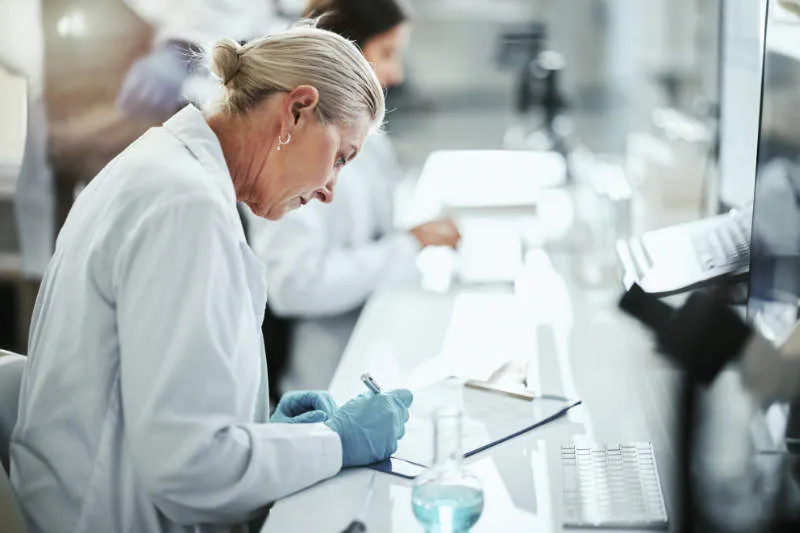 A female scientist in a lab coat reviews notes at a laboratory bench.