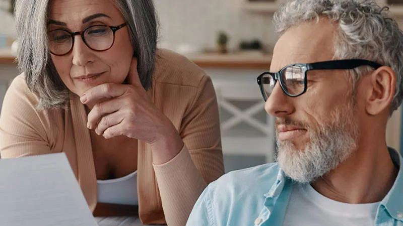 An older couple with glasses examines a document together at home. The woman rests her cheek on her hand, looking thoughtful, while the man smiles slightly. They are in a kitchen or dining area.