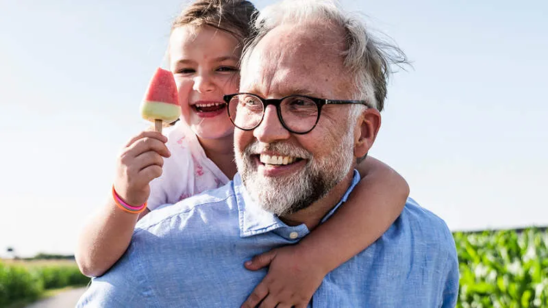 A smiling older man with glasses and a white beard carries a happy little girl on his shoulders. She holds a watermelon popsicle and hugs him, both enjoying a sunny day outdoors near a green field.