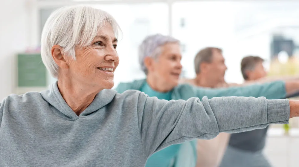 A smiling older woman with short white hair participates in an exercise class, stretching her arms forward while other seniors exercise in the background.